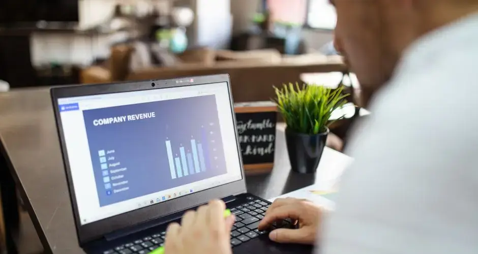 Man using a laptop to analyze company revenue data with graph charts in office setting.