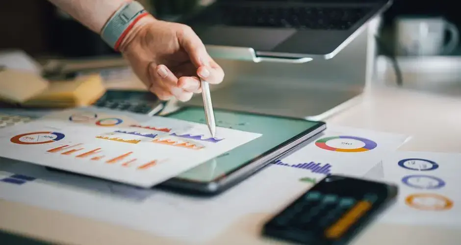 Hand using a tablet and charts on desk to analyze business data. Ideal for finance, business, and technology themes.