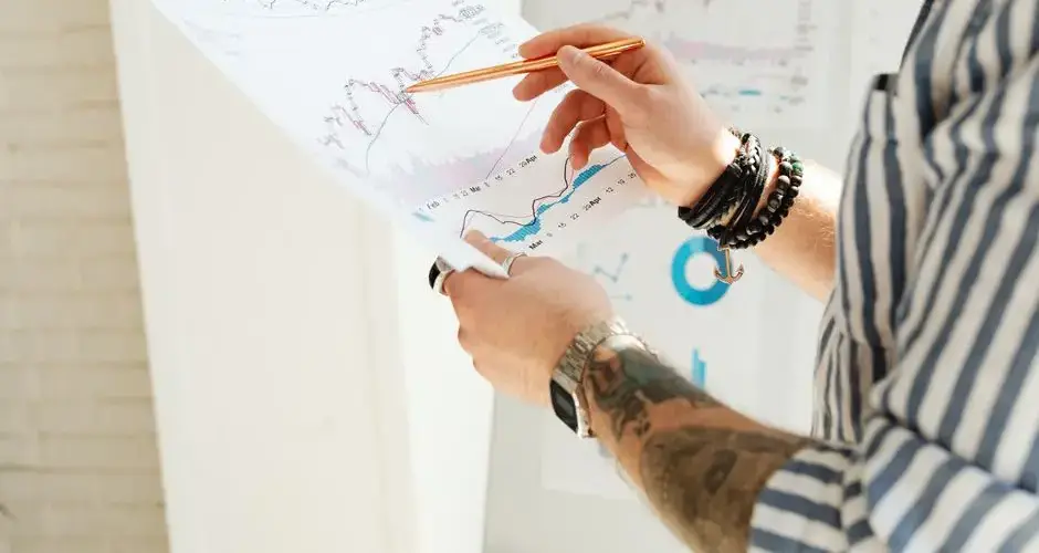 A man analyzing stock market charts with a pen, holding a paper report indoors.