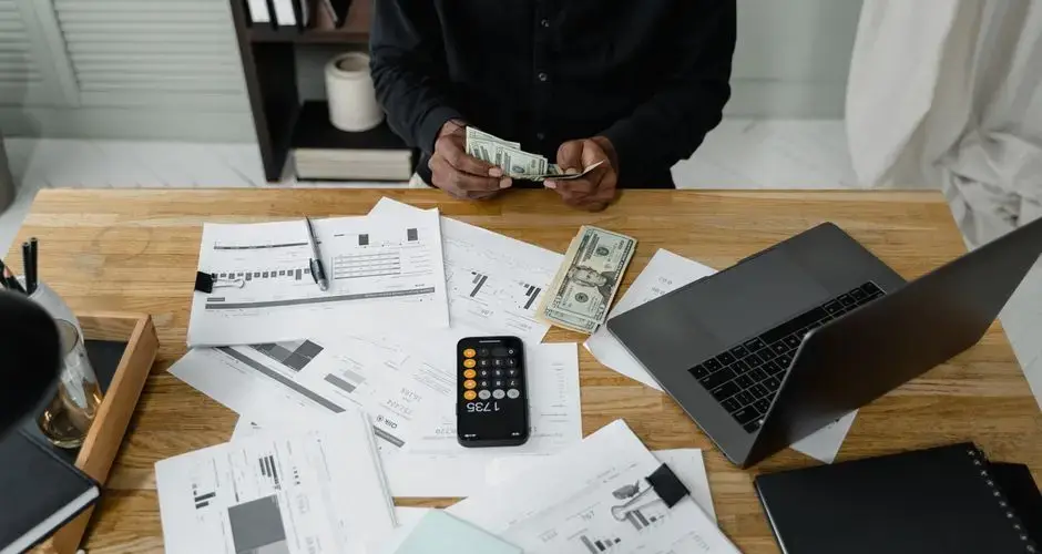 A person counts dollar bills at a desk with a laptop, papers, and calculator.