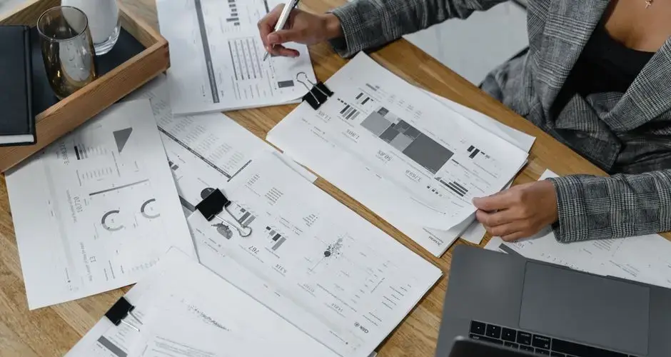 Woman analyzing financial data and reports on a wooden desk with a laptop, showcasing analytics in a workspace.