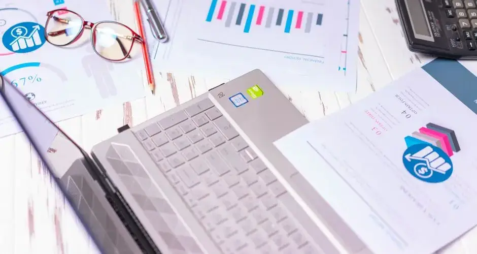 A cluttered desk with a laptop, financial reports, calculator, and glasses indicating a busy work environment.