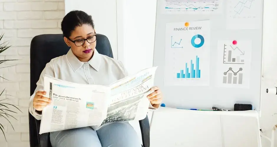 A woman reads a newspaper with stock charts displayed in an office setting.