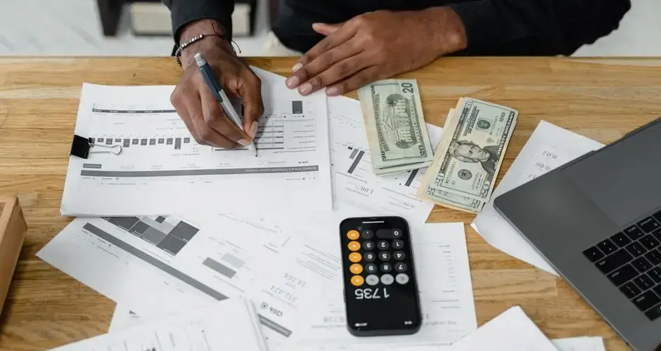 Man working on financial reports with calculator, money, and laptop on a desk.