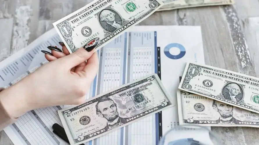 Close-up of a hand holding a dollar bill over financial documents and charts on a wooden table.