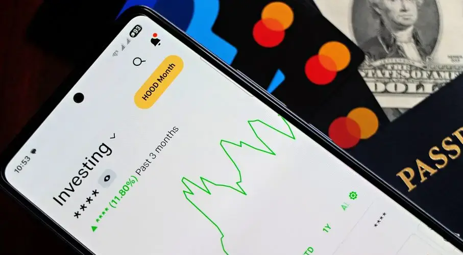 A smartphone showing an investment app with green growth indicators, surrounded by credit cards, US dollars, and a passport.