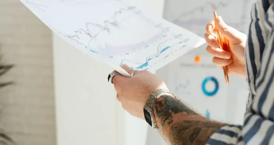 Close-up of a tattooed man's hands holding financial charts, with focus on data analysis and stock market trends.