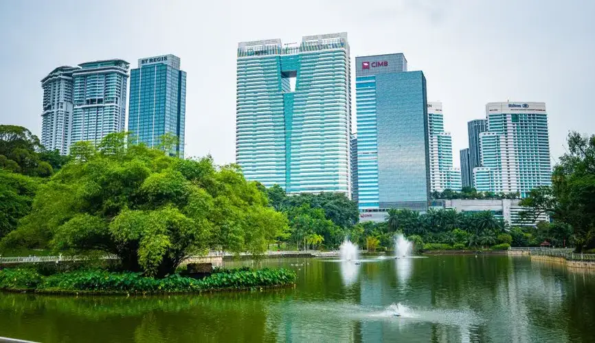 Modern skyline of Kuala Lumpur with lush park and water fountain in the foreground.
