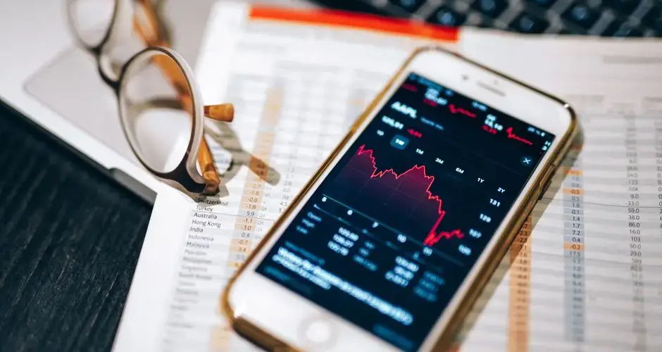 Smartphone displaying stock market chart on a financial spreadsheet with eyeglasses.