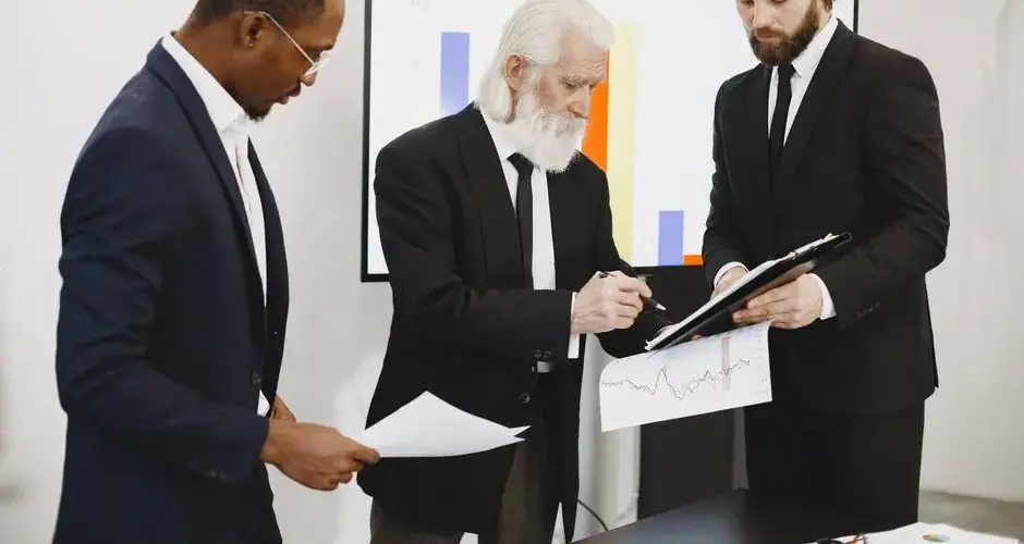 Three businessmen in suits reviewing reports and graphs during an office meeting.