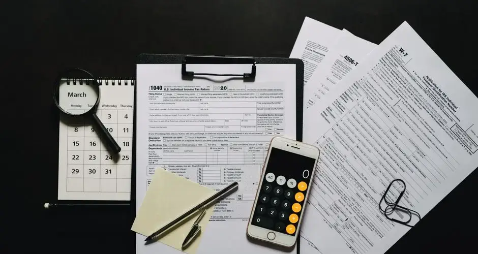 Top view of tax documents, calculator, magnifying glass, and calendar on a black surface.