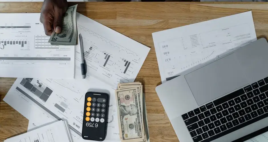 Overhead view of a busy workspace with cash, financial documents, and a laptop.