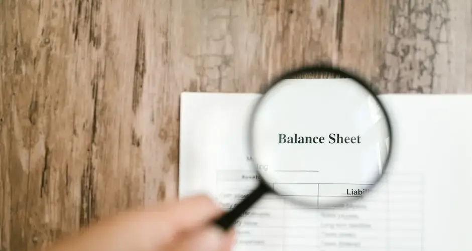 Close-up of a balance sheet document on wooden surface with a magnifying glass held by a hand.