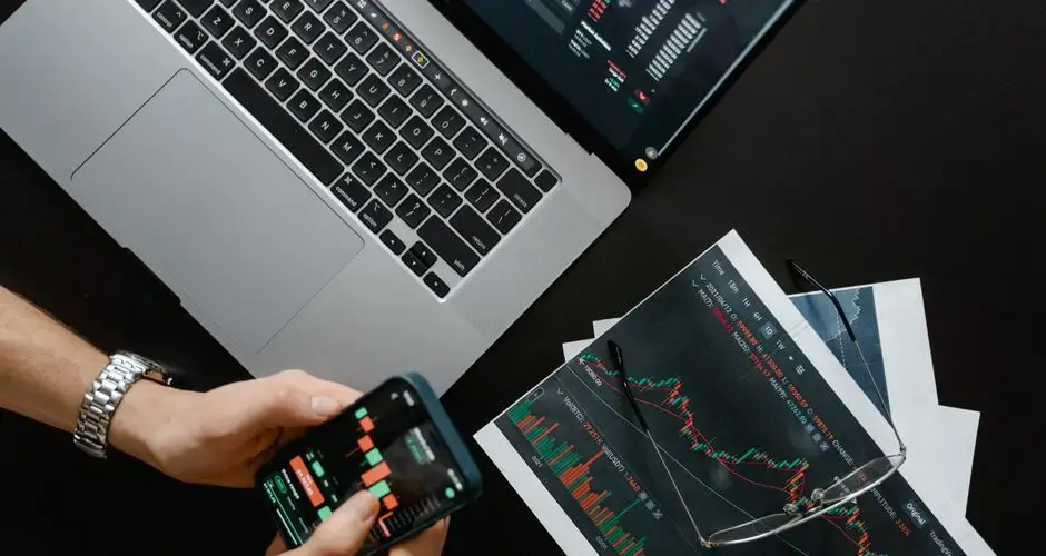 Hands using smartphone beside laptop with stock charts, showcasing digital trading.