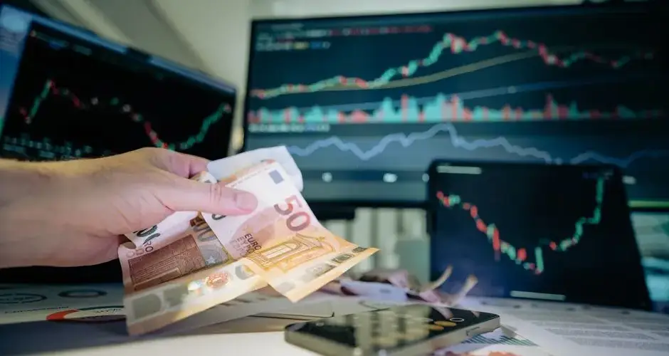 Business professional counting Euro notes with financial charts on screens, indicating investment strategy.