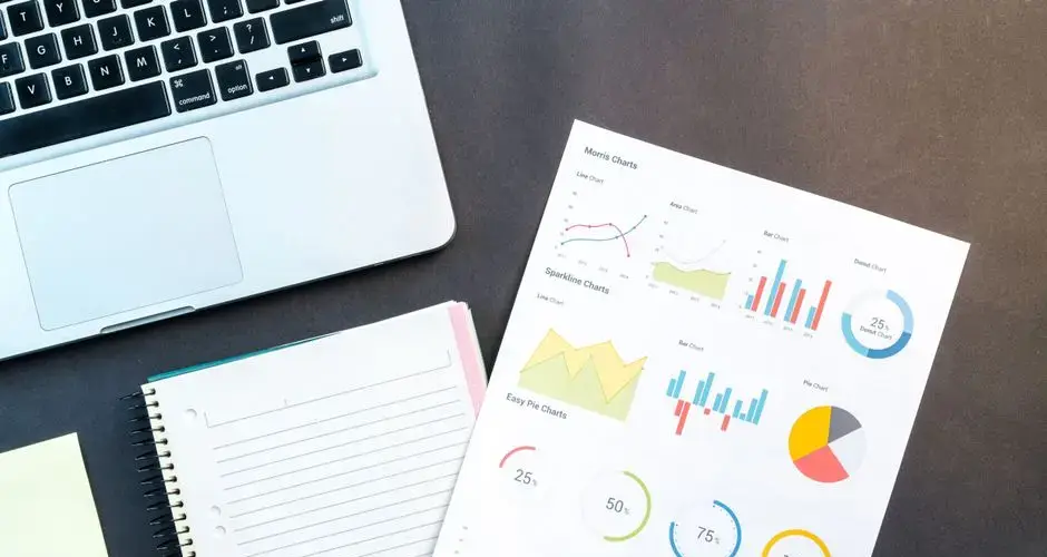 Flatlay of business report with colorful charts, a notebook, and a laptop for data analysis on a desk.