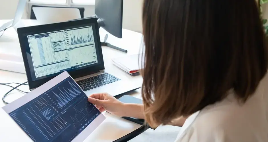 A professional woman analyzes financial charts on a laptop and paper for business insights.