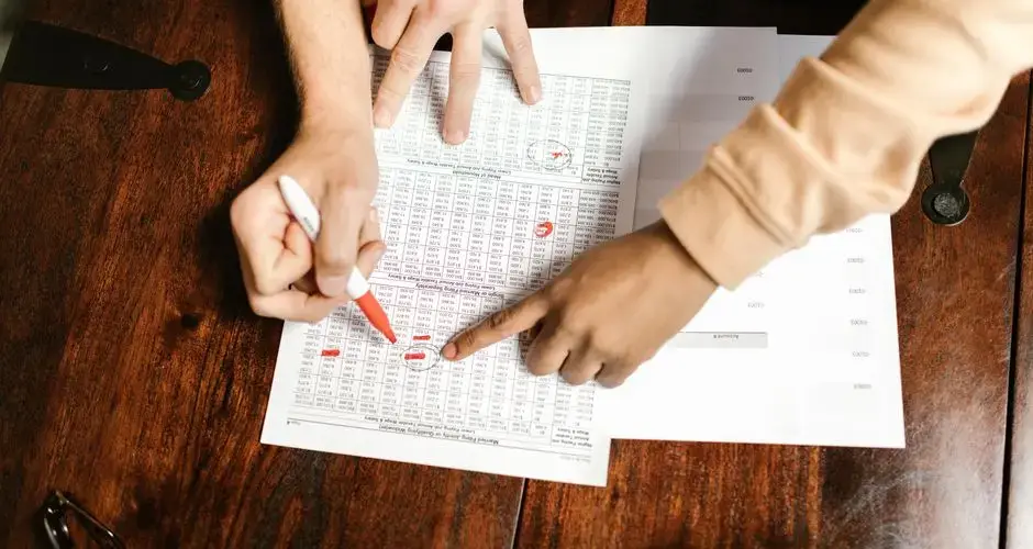Two hands reviewing and marking financial documents with a red pen on a wooden table, top view.