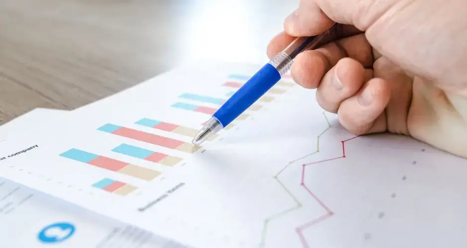 A close-up of a hand with a pen analyzing data on colorful bar and line charts on paper.