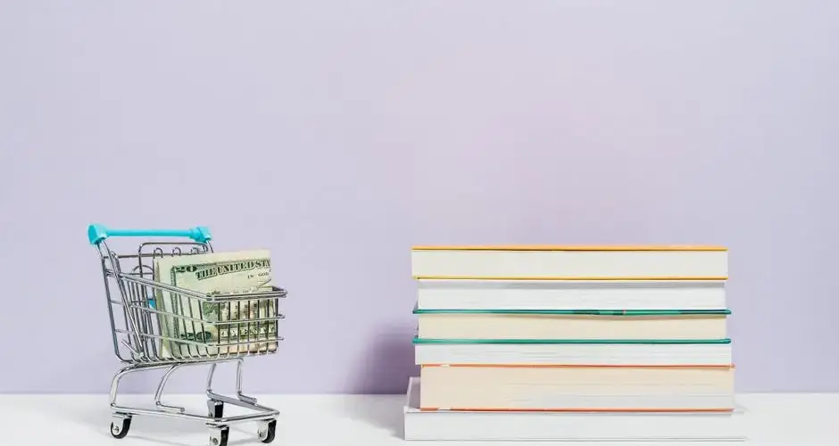 A shopping cart with dollar bills next to a stack of textbooks on a light background.