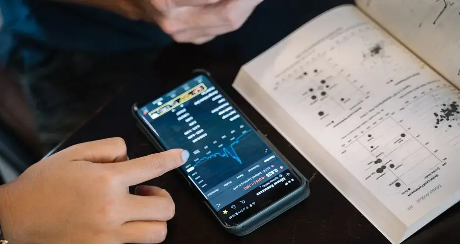 Hand pointing at smartphone displaying financial data, with open book nearby.