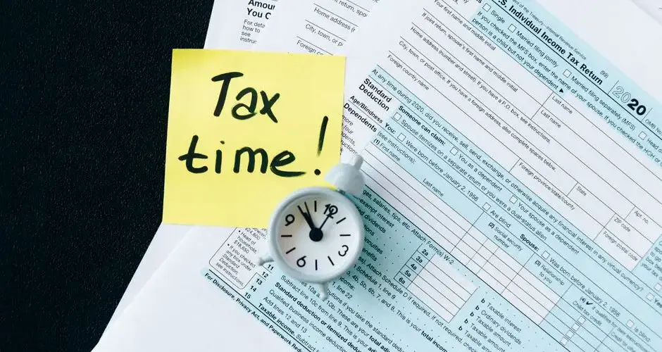 A stack of tax forms with a clock and yellow sticky note saying 'Tax time!' indicating urgency.