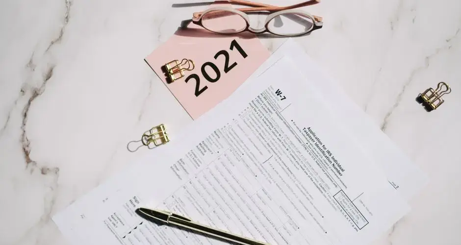 Close-up of W-7 tax forms with glasses and pen on a marble desk, ideal for finance concepts.