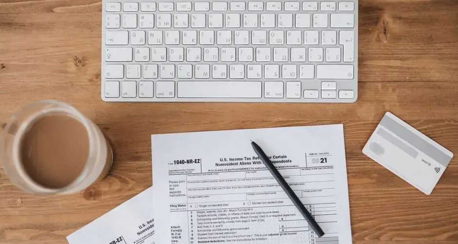 Top view of tax forms, keyboard, pen, and coffee on a wooden desk.