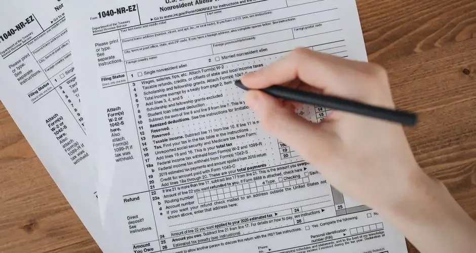 Person filling out U.S. tax form 1040-NR-EZ with a pen on a wooden table.
