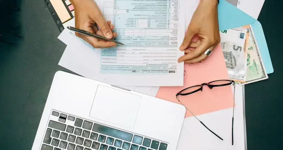 Hands writing on tax documents with laptop, glasses, and currency on desk.
