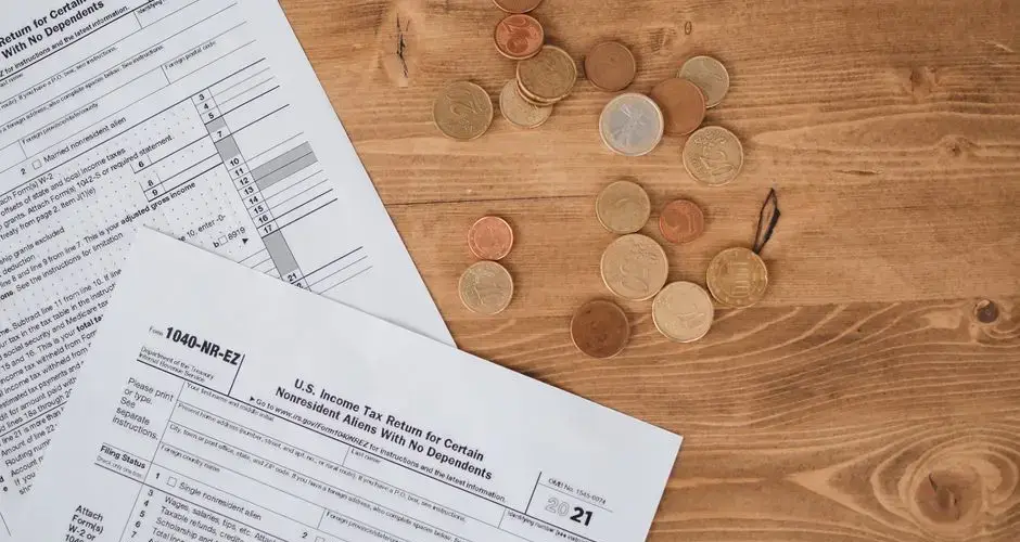 Flat lay of tax forms and scattered coins on a wooden table, illustrating finance and taxation concepts.
