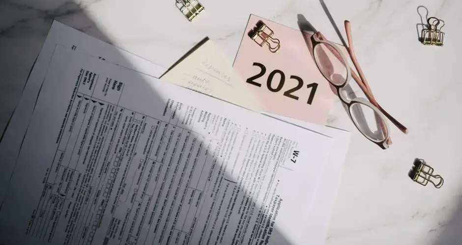 Close-up of tax documents, glasses, and a note labeled 2021 on a marble desk.