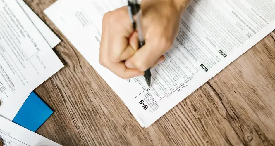 Hand writing on a W-9 tax form using a ballpoint pen on wooden desk.