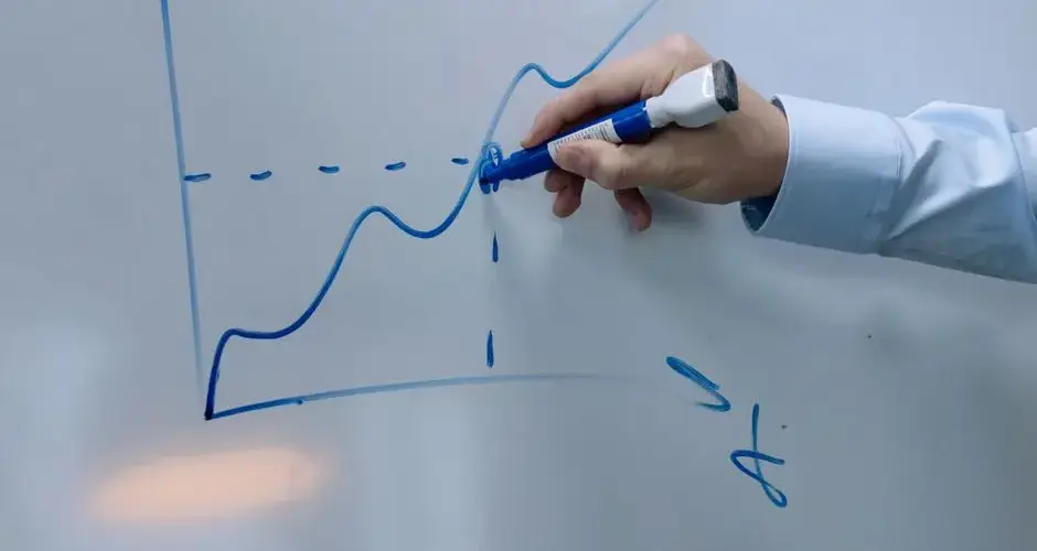 Hand drawing an upward graph on a whiteboard using a blue marker being held by a person in a formal shirt.