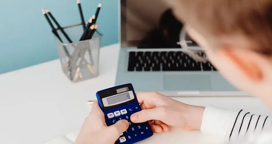 Close-up of hands using a blue calculator at a desk with a laptop and stationery.
