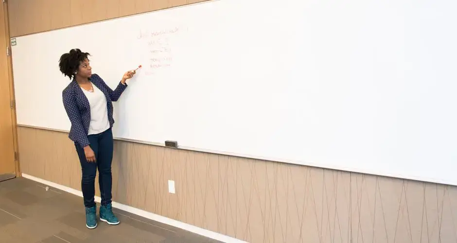 Black female educator explaining concepts on whiteboard in conference room setting.