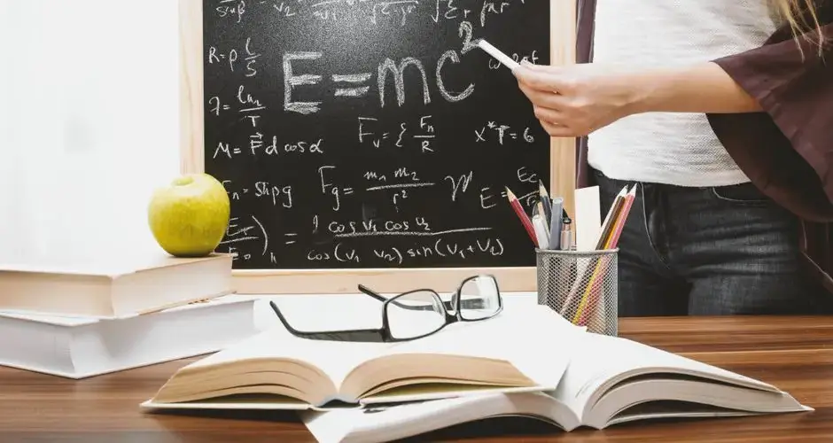 Woman writing physics equations on a blackboard with books and an apple on the desk.