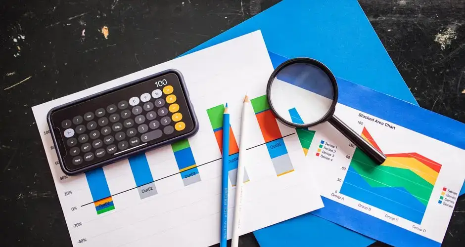 Top view of financial charts with a smartphone calculator, magnifying glass, and pencils on a desk.