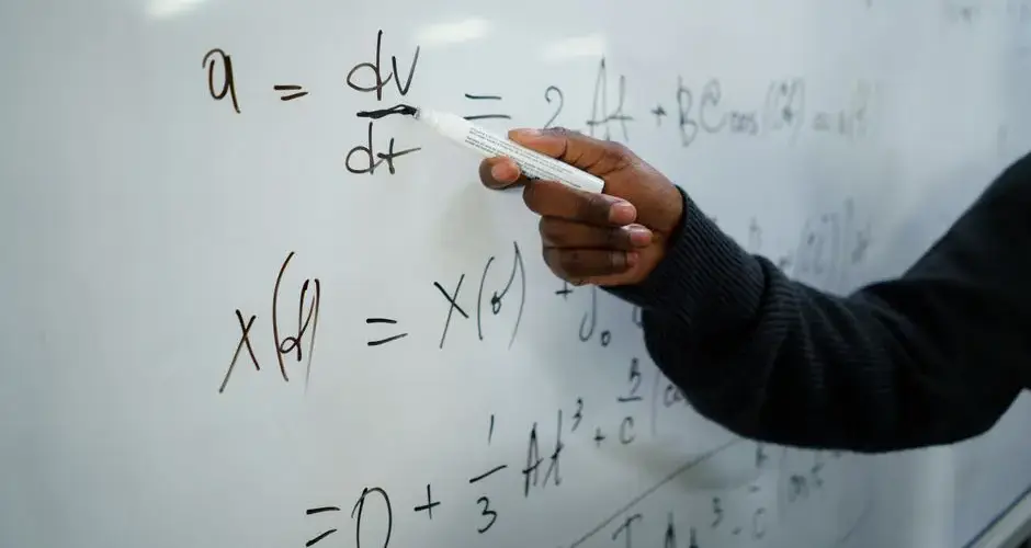 Close-up of hand writing equations on a whiteboard in a classroom setting.