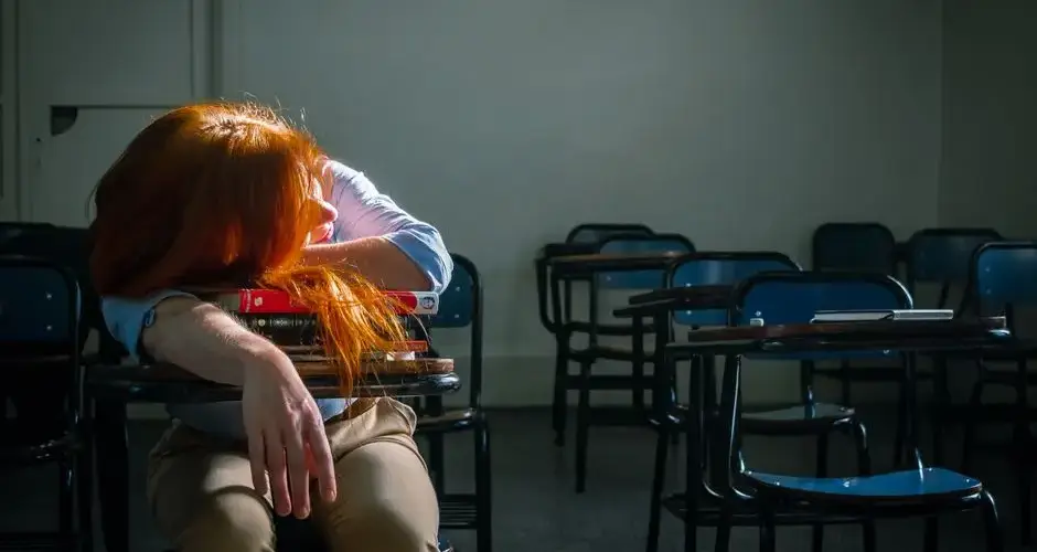 A tired student with red hair sleeps in an empty classroom under soft natural light.