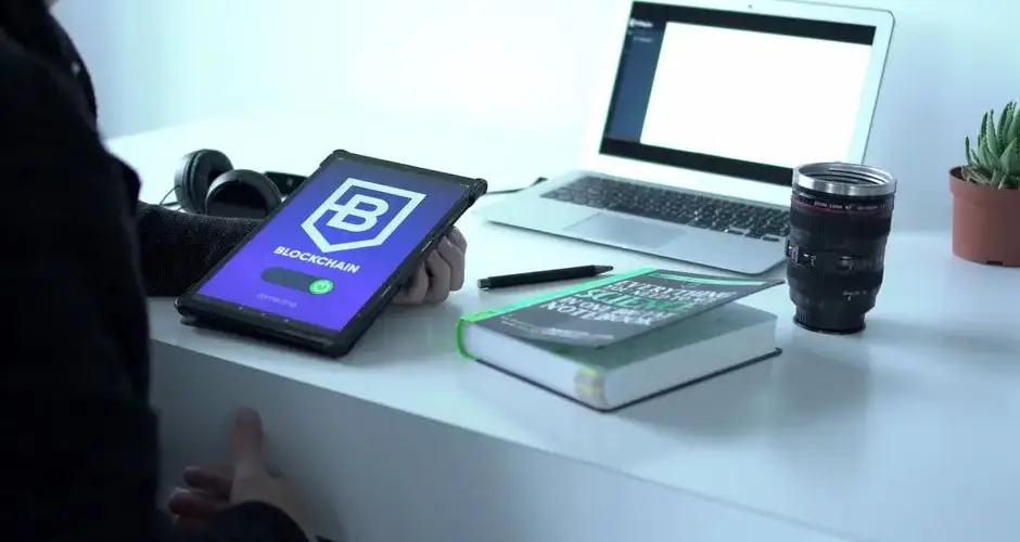 A desk with tablet displaying blockchain, laptop, book, and camera lens, symbolizing tech and innovation.