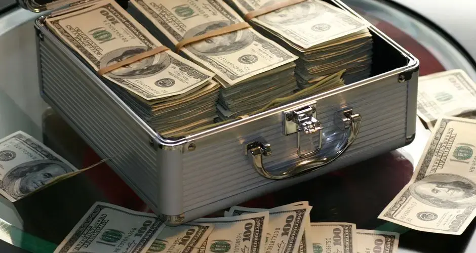 Open briefcase filled with stacks of hundred dollar bills on a glass table, representing wealth.
