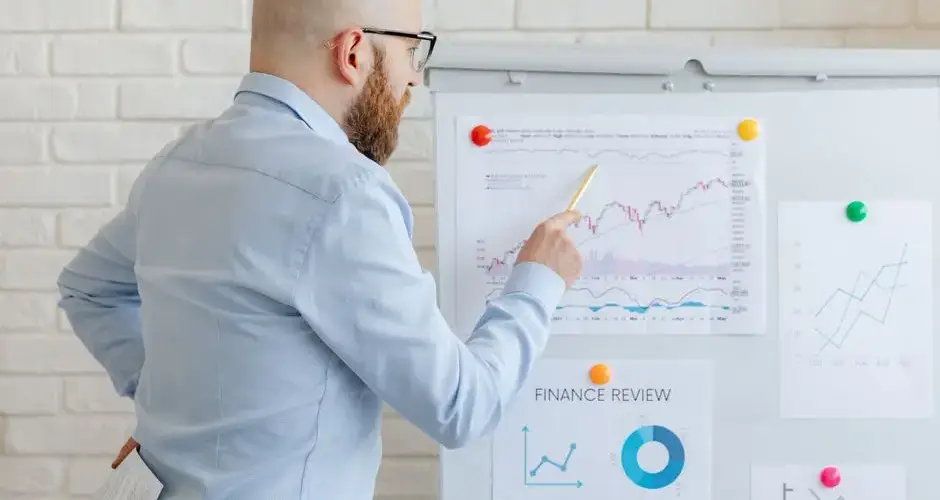 Bald businessman in office analyzes graphs on whiteboard, indicating finance trends.
