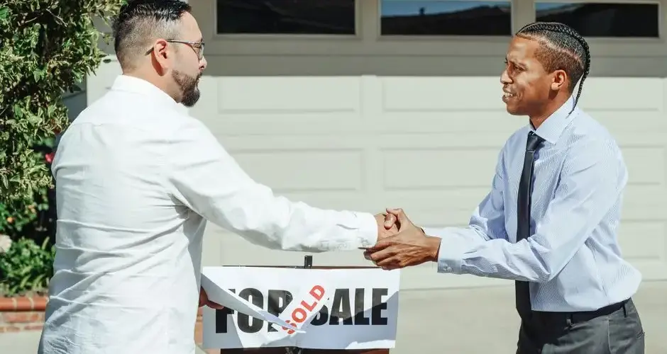Two men shaking hands in front of a sold house sign, sealing a real estate transaction.