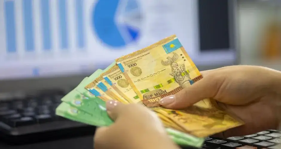 Hands holding Kazakhstan tenge bills with financial chart and graphs on computer screen.