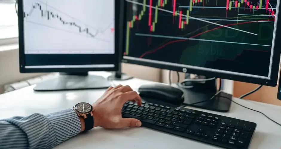 A businessperson using dual monitors to analyze stock market trends with charts and graphs.