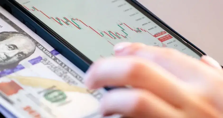 Close-up of a smartphone displaying stock market data over a dollar bill on a desk.