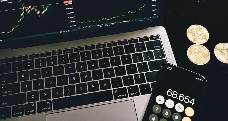 A laptop with trading charts, smartphone calculator, and bitcoin coins depicting cryptocurrency trading.