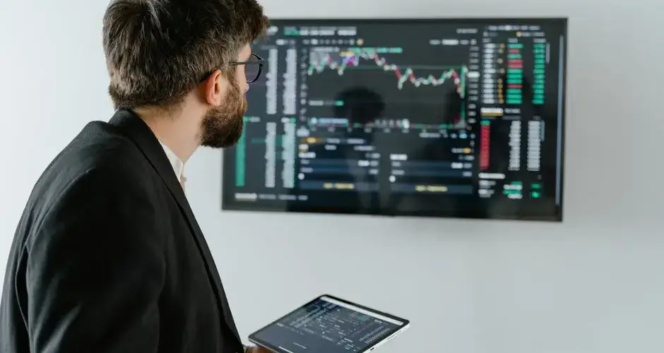A businessman examines stock market data displayed on a monitor, holding a tablet.