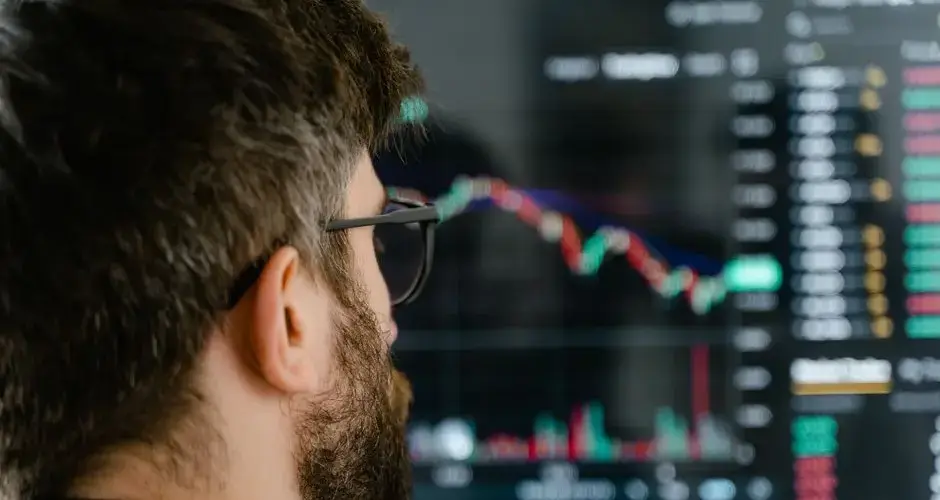 A bearded man with glasses examines trading charts on a computer screen, indicating financial analysis.